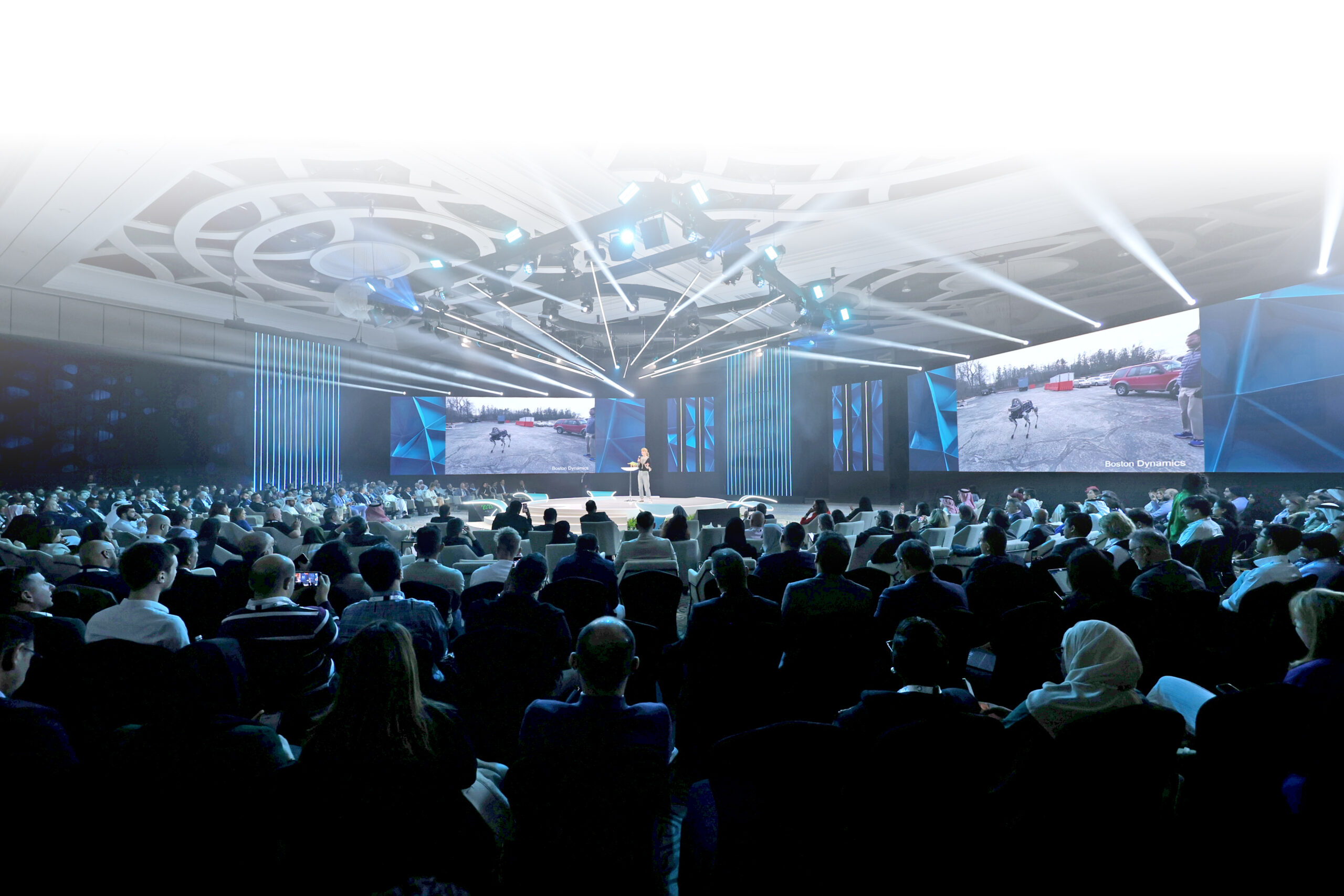 A speaker on stage in a packed summit auditorium, with Boston Dynamics robotics footage shown on screens either side
