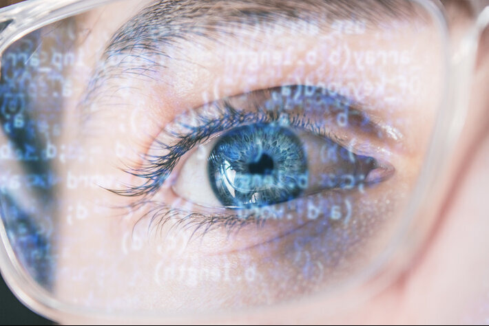 Closeup shot of hacker's blue eye wearing optical glasses with data code reflecting on the glass